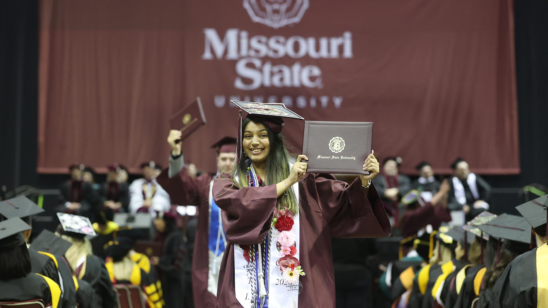 A brand new #BearGrad happily holds up her diploma cover while walking at commencement.