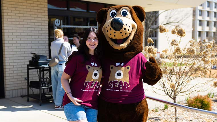 A student with Boomer outside of the bookstore. Both are wearing Maroon Be A Bear t-shirts.