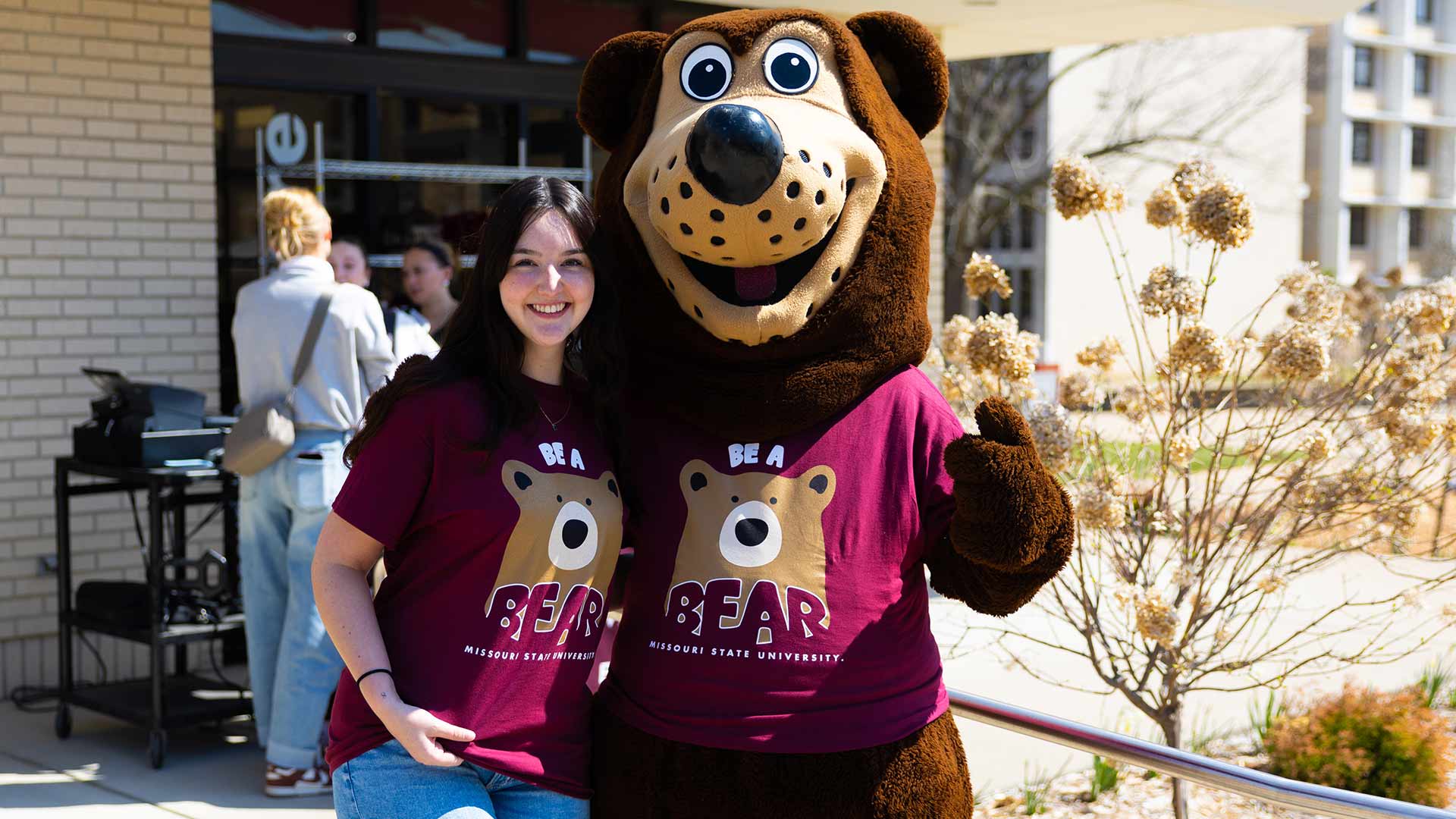 A student with Boomer outside of the bookstore. Both are wearing Maroon Be A Bear t-shirts.