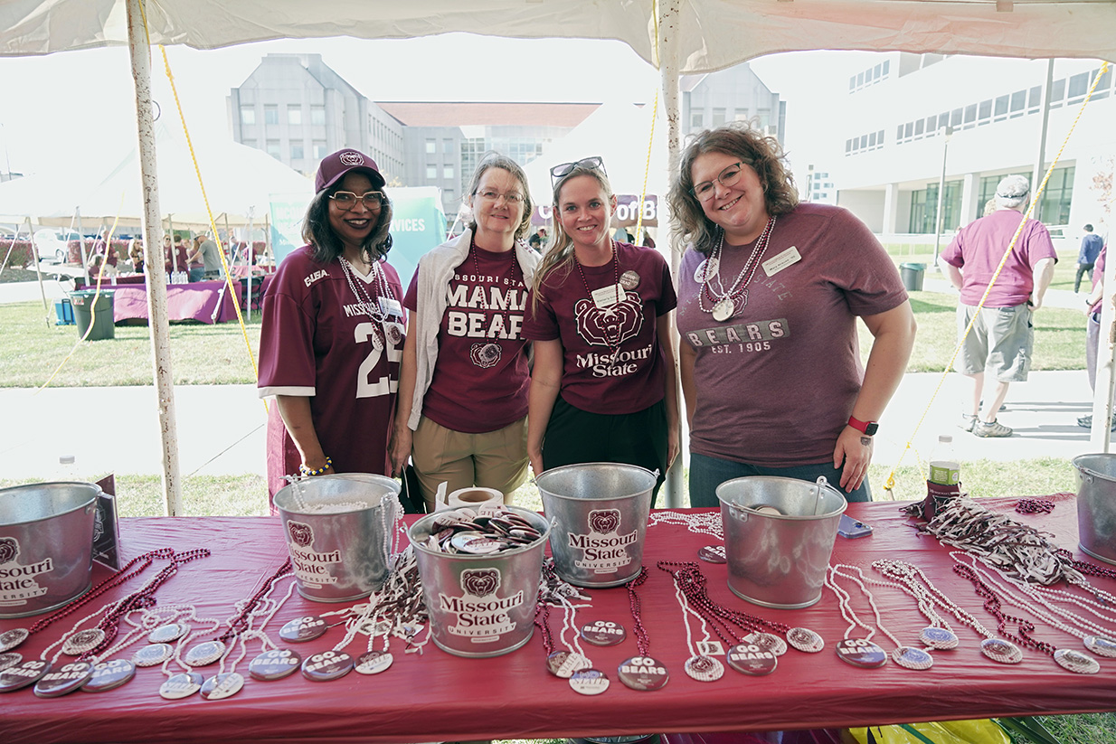Photo of four women standing together behind a table of Missouri State beads and pom poms. Everyone is smiling and wearing BearWear.