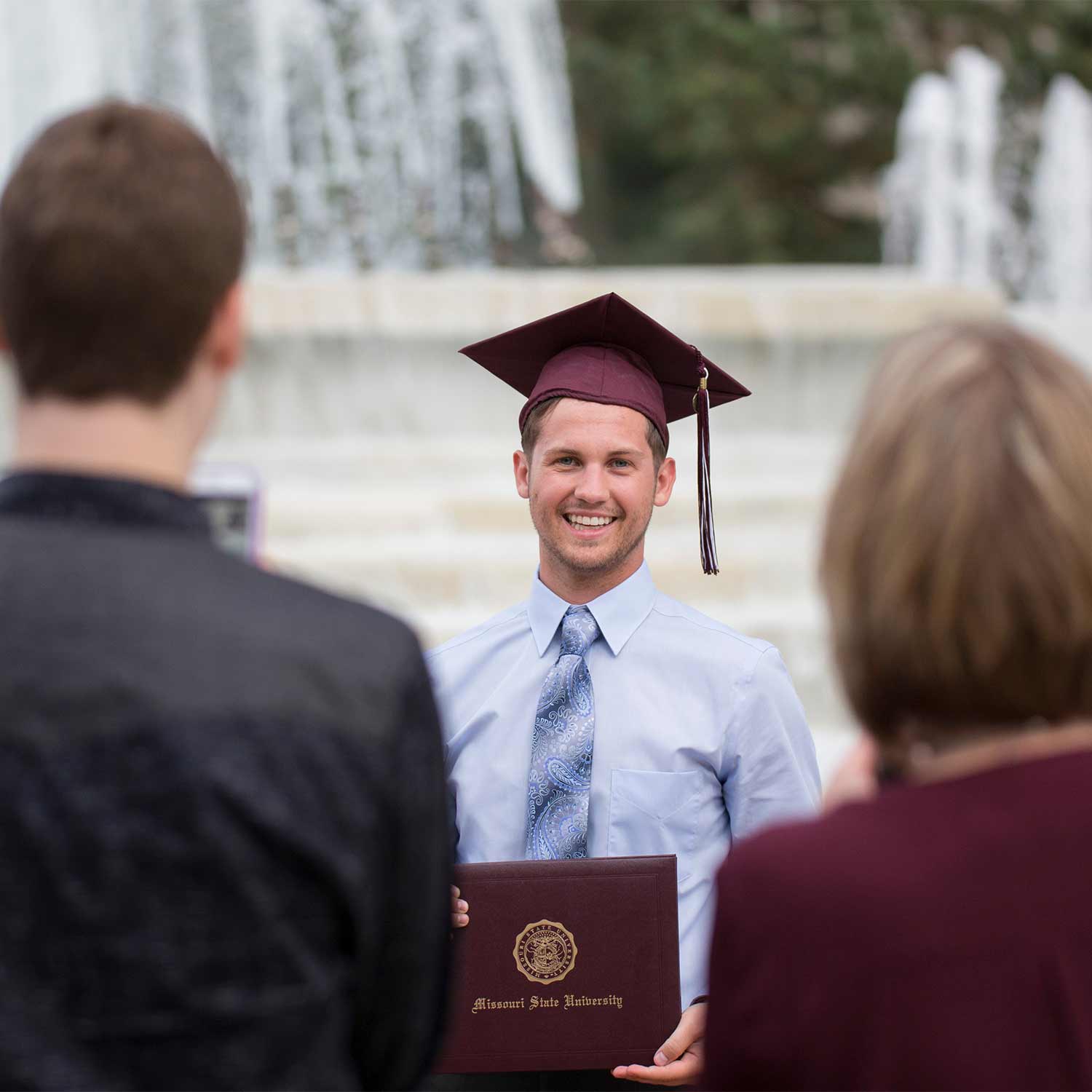 A #BearGrad wearing a maroon mortar and holding up his diploma cover poses for a photo in front of the campus fountain.