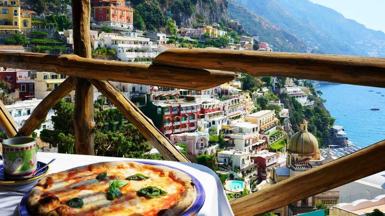 A view of the Amalfi Coast and community homes from a restaurant in Italy. A plate of food and teacup can be seen in the foreground.